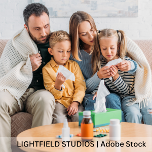 A family of four huddled together with a blanket on the couch while feeling sick. Liquid medicine, pills, and tissues out on the coffee table.