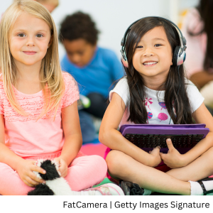 Two young girls smiling and sitting cross-legged on the floor with one girl holding a stuffed animal and the other with headphones on her head and holding an ipad with a keyboard attached it.