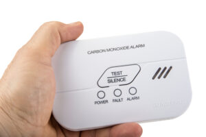 Close-up of a hand holding a white carbon monoxide alarm device. The alarm has three buttons labeled ‘TEST,’ ‘SILENCE,’ and indicators for ‘POWER,’ ‘FAULT,’ and ‘ALARM.’ Text on the device reads ‘CARBON MONOXIDE ALARM’ and ‘DO NOT PAINT.’