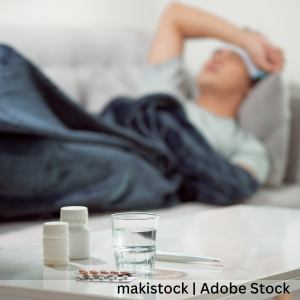 A person lying on a couch under a blanket, with medicine bottles, a blister pack of pills, a glass of water, and a thermometer placed on a table in the foreground.