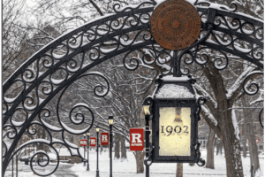 Snow-covered iron gate at Rutgers University with a lantern marked “1902” and campus banners in the background.