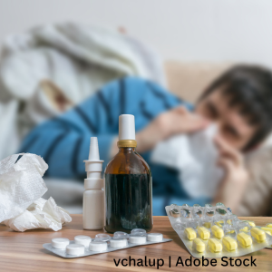 Close-up of cold and flu medications on a table, including blister packs of pills, a nasal spray, and a brown glass bottle, with crumpled tissues nearby. In the blurred background, a person is lying down and holding a tissue to their nose, suggesting illness.