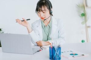 A healthcare professional wearing a white coat and headset sits at a desk, speaking with a caller on the phone. 