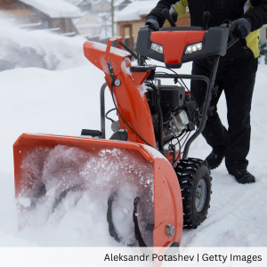 A person operating an orange snowblower outdoors during heavy snowfall, clearing snow from a driveway or walkway.