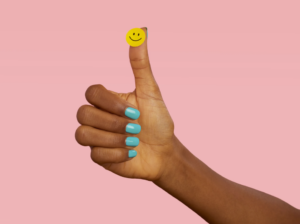 A hand giving a thumbs‑up gesture, with a small yellow smiley face sticker on the thumb, against a pink background.