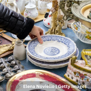 A hand reaching to pick up a stack of blue‑and‑white patterned ceramic plates on a table filled with assorted vintage dishware and decorative items.