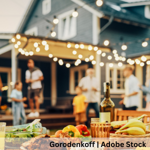 Backyard gathering with table of food in foreground and people socializing on a porch under string lights at dusk.